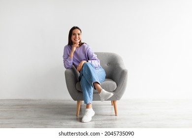 Full Length Portrait Of Happy Young Woman In Casual Wear Smiling At Camera, Sitting In Comfy Armchair Against White Studio Wall. Lovely Millennial Lady Posing, Relaxing Indoors