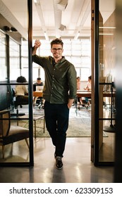 Full Length Portrait Of Happy Young Businessman Standing In Doorway Of Office. Caucasian Male Executive In Office With People Working In Background.