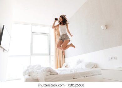 Full length portrait of a happy young woman in earphones listening to music and jumping on bed indoors