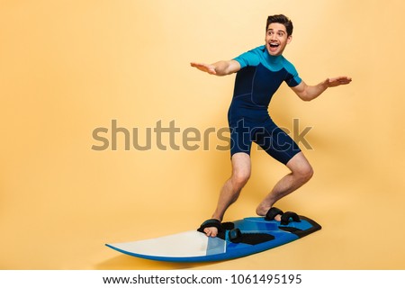 Full length portrait of a happy young man dressed in swimsuit surfing on a board isolated over yellow background