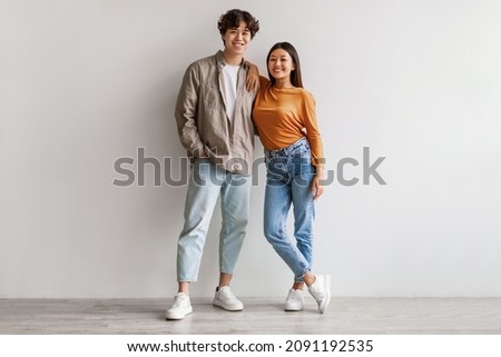 Full length portrait of happy young Asian couple in casual clothes hugging and looking at camera against white studio wall. Cheerful millennial spouses posing and smiling together
