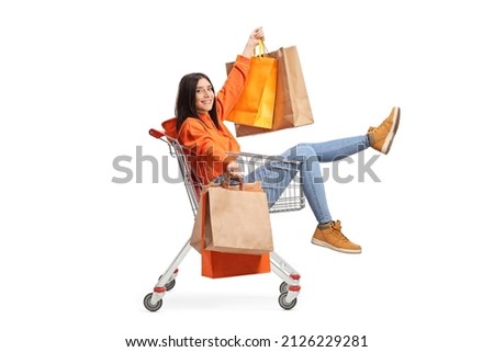 Full length portrait of a happy young female sitting inside a shopping cart and holding shopping bags isolated on white background