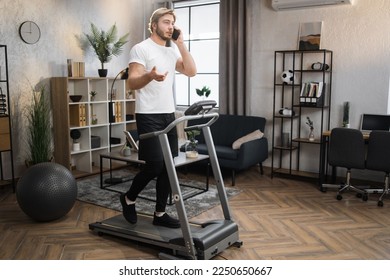 Full length portrait of healthy sporty man, talking on his smartphone while doing cardio training at home. Blond bearded male in white t-shirt jogging on treadmill. - Powered by Shutterstock