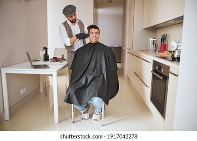 Full Length Portrait Of Indian Neat Man In Black Peignoir Sitting On The Chair While Barber Making New Styling With Hair In The Home Kitchen