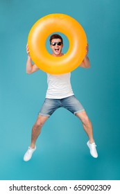 Full length portrait of a joyful happy guy in sunglasses jumping and looking through inflatable ring isolated over blue background