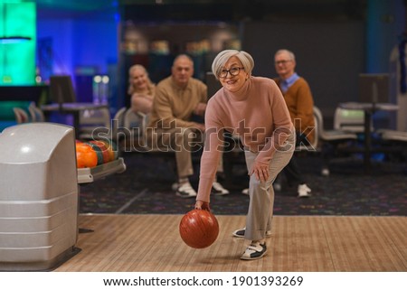 Full length portrait of joyful senior woman playing bowling with group of friends in background, copy space