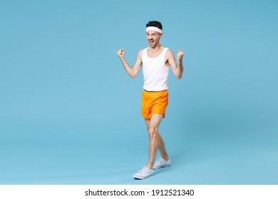 Full Length Portrait Joyful Young Sporty Fitness Man With Skinny Body Sportsman In White Headband Shirt Shorts Doing Winner Gesture Isolated On Blue Background. Workout Gym Sport Motivation Concept