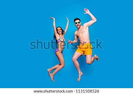 Full length portrait of a joyful young woman and happy man in sunglasses, dressed in swimsuit, jumping and putting hands up over blue background