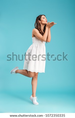 Full length portrait of a lovely young girl in summer dress sending kiss while standing isolated over blue background