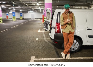 Full length portrait of male delivery worker standing by van at parking lot and using smartphone, copy space - Powered by Shutterstock