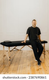 Full length portrait of a male masseur standing behind a professional massage bed