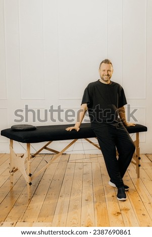 Full length portrait of a male masseur standing behind a professional massage bed