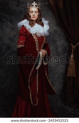 Full length portrait of medieval queen in red dress with white collar and crown on dark gray background.