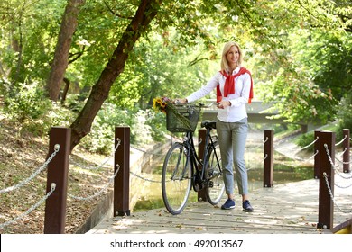 Full Length Portrait Of Middle Aged Woman Pushing Her Bike In The Park While Commuting In The City.