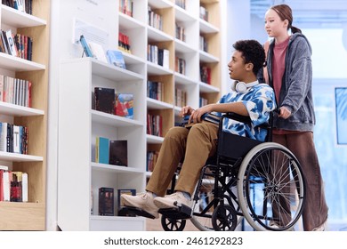 Full length portrait of Middle Eastern boy using wheelchair choosing books on shelves in school library with friend assisting copy space - Powered by Shutterstock