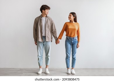 Full length portrait of millennial Asian couple smiling and looking at each other, holding hands, standing against white studio wall. Affectionate young man and woman being in love - Powered by Shutterstock