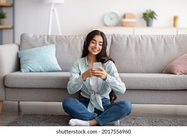 Full length portrait of millennial Indian lady in headphones using cellphone, listening to music, sitting on floor at home. Cheerful young woman picking favorite playlist on mobile device