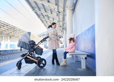 Full length portrait of a mother with children waiting train on the railway station platform. Pretty woman commuter pushing baby pram and talking to her daughter while travelling together by train