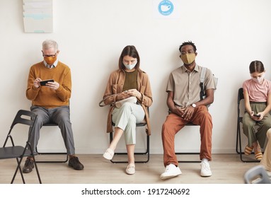 Full Length Portrait Of Multi Ethnic Group Of People Wearing Masks And Sitting In Row On Chairs While Waiting In Line At Clinic, Copy Space