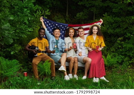 Full length portrait of multi-ethnic group of people holding American flag while sitting on bench in forest and enjoying Summer vacation, copy space