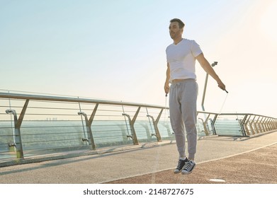 Full length portrait of a muscular build European middle aged man, fit athlete doing jumping exercises, cardio training with skipping rope on the urban city bridge early in the morning on a summer day - Powered by Shutterstock