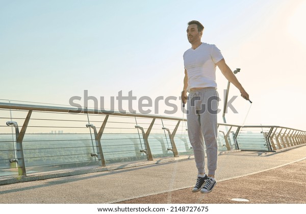 Full length portrait of a muscular build European\
middle aged man, fit athlete doing jumping exercises, cardio\
training with skipping rope on the urban city bridge early in the\
morning on a summer day