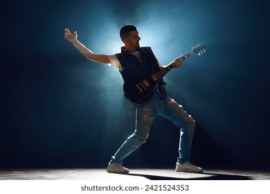 Full length portrait of music artist in ripped jeans and vest bending strings on guitar under spotlights on stage with smoke. Concept of Rock-n-roll, music and dance, festivals and concerts, culture. - Powered by Shutterstock