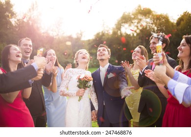 Full Length Portrait Of Newlywed Couple And Their Friends At The Wedding Party Showered With Confetti In Green Sunny Park