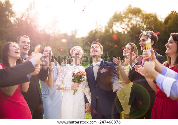 Full\
length portrait of newlywed couple and their friends at the wedding\
party showered with confetti in green sunny\
park