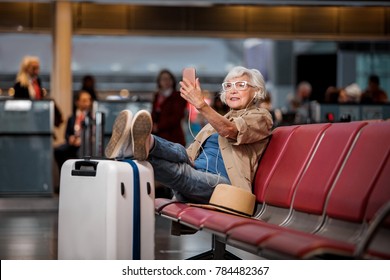 Full Length Portrait Of Old Lady Is Sitting On Bench At Airport Lounge. She Is Having Video Call Using Smartphone And Earphones While Putting Legs On Suitcase And Looking At Screen Of Gadget