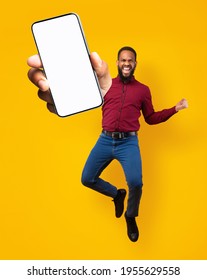 Full length portrait on joyful black man jumping, gesturing YES and showing cellphone with empty space for mobile app on white screen, orange studio background. Creative collage