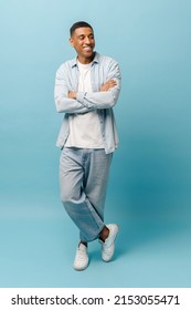 Full length portrait on stylish African-American hipster guy in denim shirt and jeans stands isolated on blue background, attractive man holding arms crossed and looking away