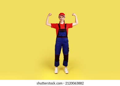 Full length portrait of optimistic woman worker standing and looking at camera with raised arms, showing her power, wearing overalls and red cap. Indoor studio shot isolated on yellow background.