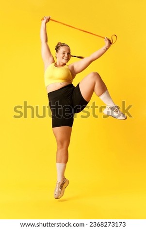 Full length portrait of plus size woman dressed sporty training, jumping with sport elastic band against over yellow background. Concept of sport, hobby, health, lifestyle, healthy eating, workout.
