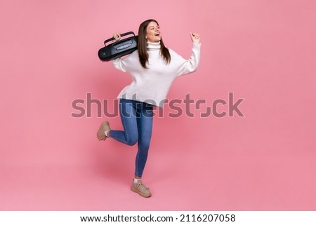 Full length portrait of positive female standing with record player, listening songs and dancing, wearing white casual style sweater. Indoor studio shot isolated on pink background.