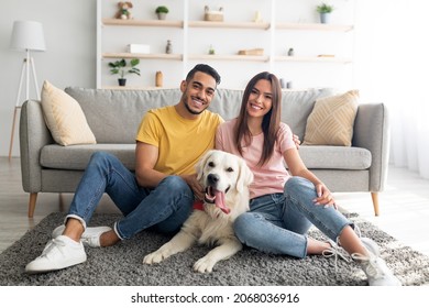 Full Length Portrait Of Positive International Couple With Their Pet Dog Sitting On Soft Carpet At Home. Happy Millennial Spouses Spending Time With Golden Retriever In Living Room