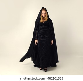 full length portrait of a pretty blonde lady wearing a gothic black dress and hooded cloak.
standing pose against a grey background.