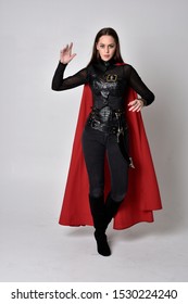 full length portrait of a pretty brunette woman wearing black leather fantasy costume with long red superhero cape. standing pose on a studio background.