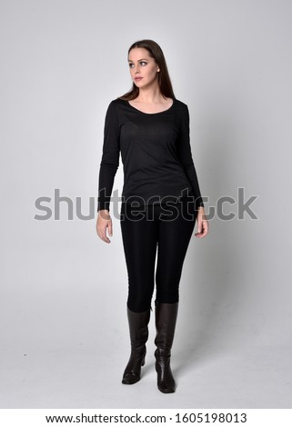 full length portrait of a pretty brunette girl wearing a black shirt leather boots. Standing pose, facing towards the camera,  on a grey studio background.