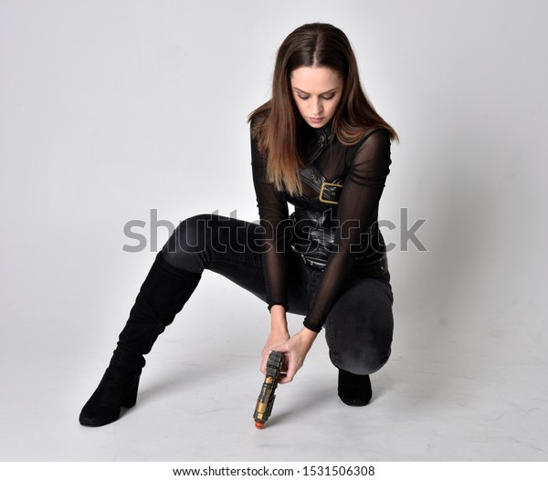 full length portrait of a pretty brunette\
woman wearing black leather fantasy costume. Crouching pose on a\
studio background.