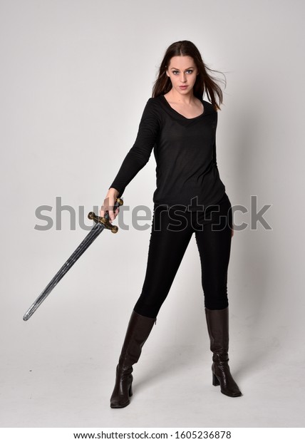 黒いシャツと革のブーツを身に着け 剣を持つ美しいブルネットの女の子の全長のポートレート グレイのスタジオ背景に立ったポーズ 剣を持つ の写真素材 今すぐ編集