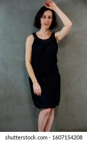 Full Length Portrait Of A Pretty Emotional Brunette Woman In Black Dress On Alternative Gray Background In Vintage Interior. Standing Right In Front Of The Camera.