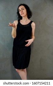 Full Length Portrait Of A Pretty Emotional Brunette Woman In Black Dress On Alternative Gray Background In Vintage Interior. Standing Right In Front Of The Camera.