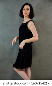 Full Length Portrait Of A Pretty Emotional Brunette Woman In Black Dress On Alternative Gray Background In Vintage Interior. Standing Right In Front Of The Camera.