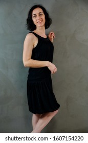 Full Length Portrait Of A Pretty Emotional Brunette Woman In Black Dress On Alternative Gray Background In Vintage Interior. Standing Right In Front Of The Camera.