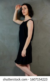 Full Length Portrait Of A Pretty Emotional Brunette Woman In Black Dress On Alternative Gray Background In Vintage Interior. Standing Right In Front Of The Camera.