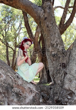  Full length portrait of pretty female model with red hair wearing green fairy dress, climbing a tree in a forest landscape.
