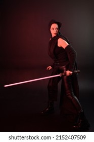 Full Length Portrait Of Pretty Redhead Female Model Wearing Black Futuristic Scifi Leather  Costume. Standing Pose With Sword, Dark Studio Background With Red Silhouette Lighting.