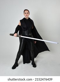 Full length portrait of pretty redhead female model wearing black futuristic scifi leather  costume. Standing pose with sword, isolated on studio background.
