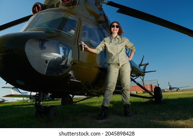 Full Length Portrait Of A Professional Female Commercial Aviation Pilot Woman In Uniform And Sunglasses Poses Happily Smiling In The Background Of A Helicopter On The Airfield. Aviation.